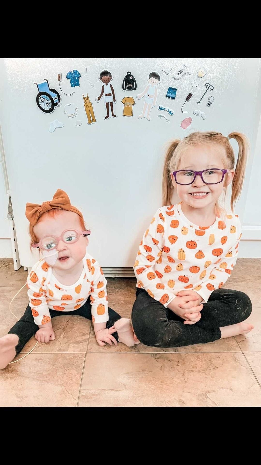 Two little children are sitting on the floor in front of a white refrigerator. Savannah, sitting on the right, is white, has glasses, blond pigtails, and is wearing a white shirt with black pants. Rosie is a baby and is sitting on the left. She has an orange bow on her head, has an NG tube inserted in her nose, is wearing glasses, and is wearing a smaller version of the same white shirt, black pants outfit as her sister. Toy magnets are on the fridge showing Bitsy Buddy feeding tube set.