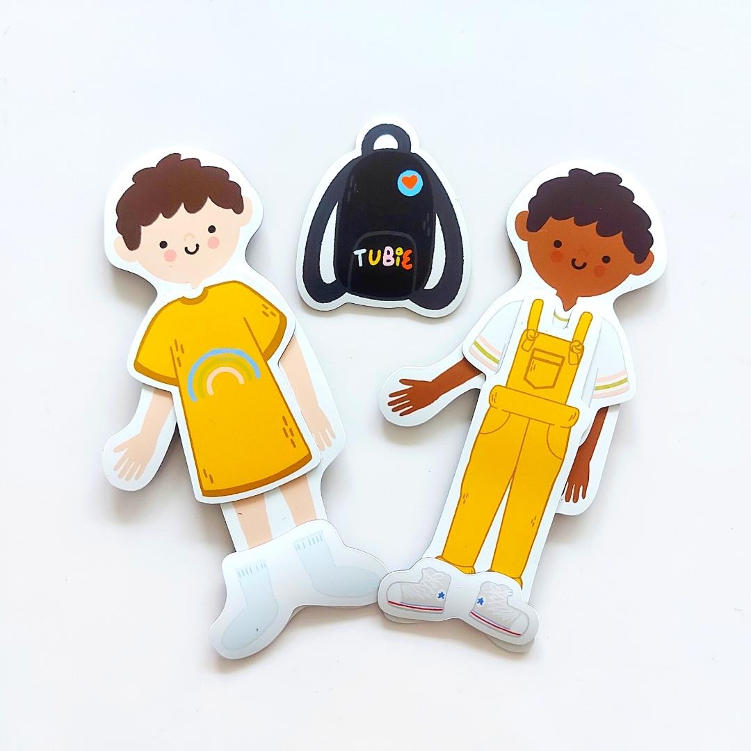 2 adorable children magnets are shown wearing a yellow rainbow dress with socks, while the other is wearing a white and green t-shirt under overalls with sneakers. Between them is a black feeding bag backpack with the word Tubie written in colourful lettering.